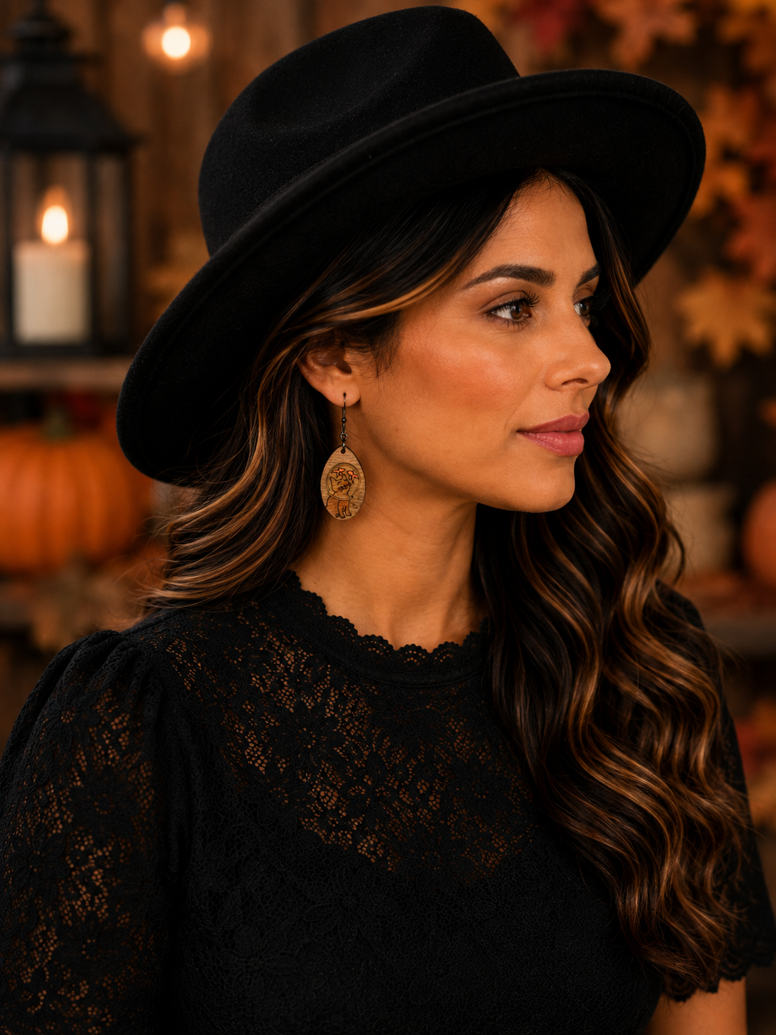 A woman in a black wide-brim hat and black lace top, wearing gold-toned dangle earrings, photographed in a warm autumn-themed setting with lanterns and pumpkins in the background.