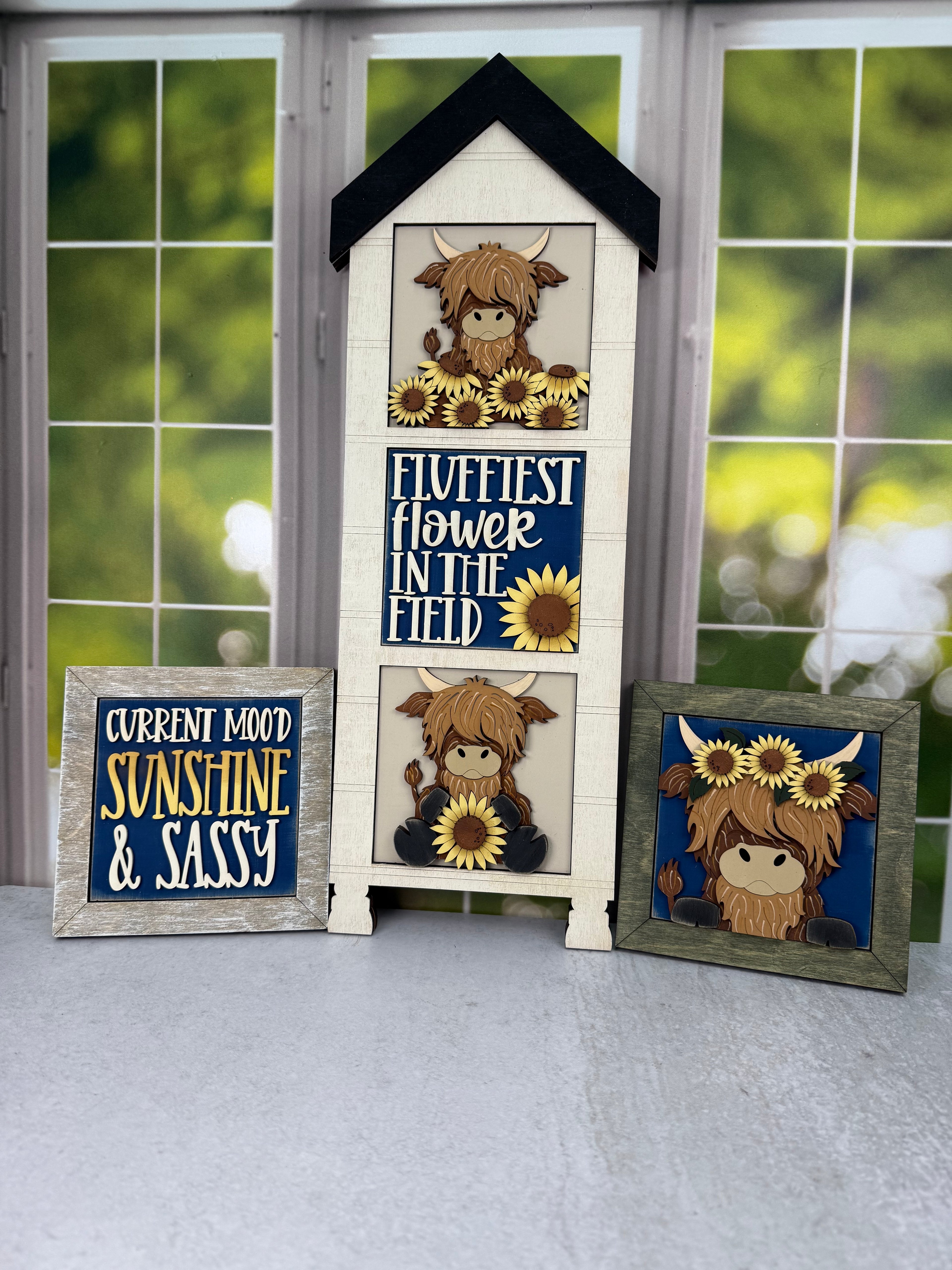 Three framed decorative signs featuring a cartoon Highland cow and sunflowers, displayed on a windowsill with a blurred outdoor garden background. The center sign is shaped like a house and reads 'Fluffiest Flower in the Field,' while the side signs read 'Current Mood Sunshine & Sassy' and show a matching cow illustration.