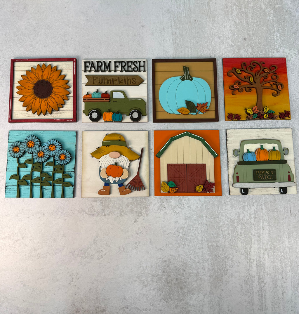 A set of eight rustic, handcrafted wooden fall-themed wall art pieces arranged in two rows of four on a textured gray background. The designs include a sunflower, a green truck carrying pumpkins with the text 'FARM FRESH Pumpkins', a light blue pumpkin, an autumn tree, blue flowers, a gnome with a rake, a barn, and a green truck with pumpkins.