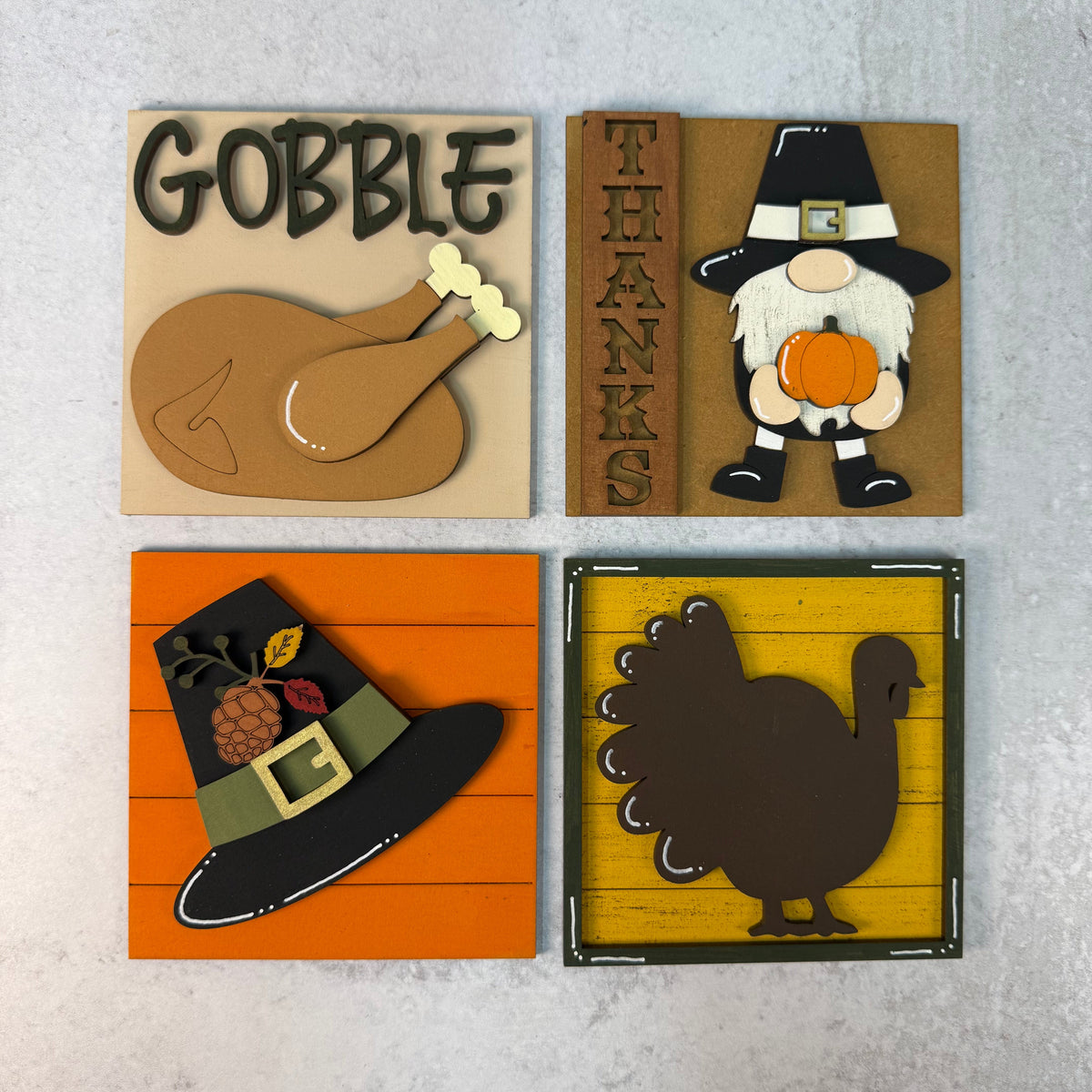 Four square Thanksgiving-themed decorative signs arranged in a 2x2 grid on a light gray textured background. The signs feature layered paper-cut designs including the word 'GOBBLE' with a turkey, a 'THANKS' sign with a pilgrim holding a pumpkin, a pilgrim hat on orange siding, and a turkey silhouette on a yellow wood-grain background.