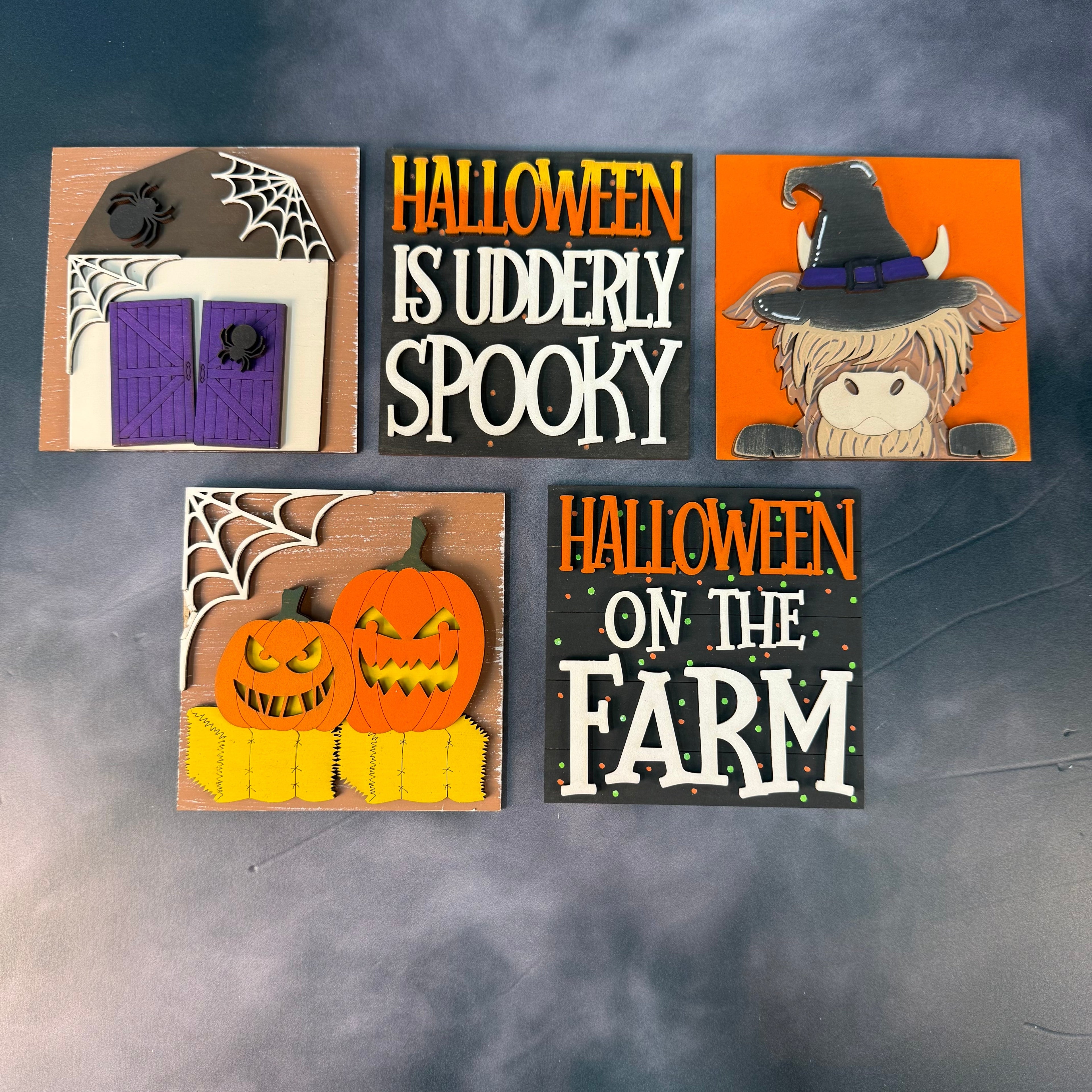 Five Halloween-themed decorative signs arranged on a smoky gray background. The signs feature designs including a haunted house with a purple door, the phrase 'HALLOWEEN IS UDDERLY SPOOKY' in orange and white, a cartoon witch on an orange background, two jack-o'-lanterns on yellow blocks, and 'HALLOWEEN ON THE FARM' in orange and white text.