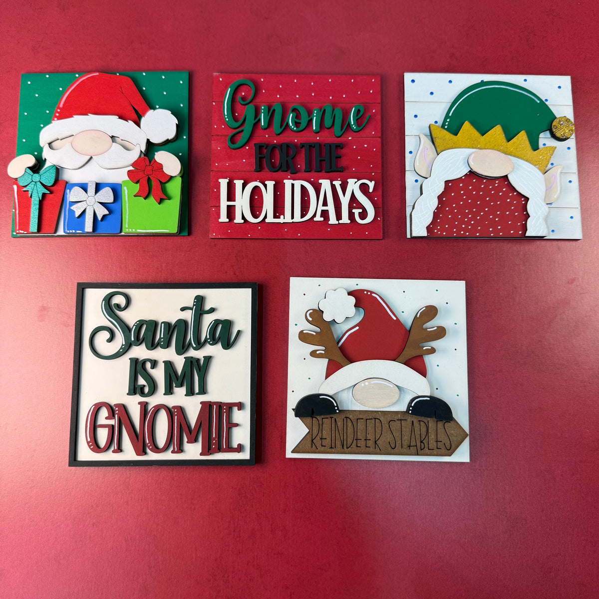 Five small square Christmas-themed decorative signs arranged on a red background. The signs feature holiday illustrations and phrases including 'Santa Is My Gnome,' 'Gnome For The Holidays,' 'Santa,' and 'MINDER STAR.