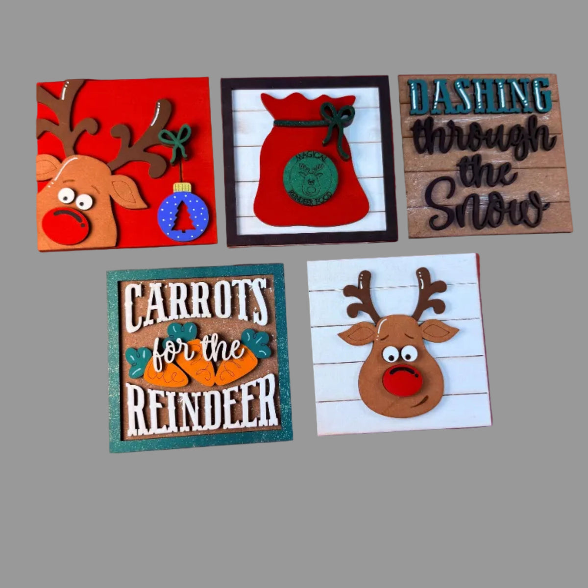 Five handmade wooden Christmas signs arranged on a red background. The signs feature holiday designs including a reindeer with antlers and a red nose, a red sack with a green ornament, the phrase 'Dashing through the Snow' on wood, 'Carrots for the Reindeer' with carrots, and a reindeer on a white shiplap-style background.