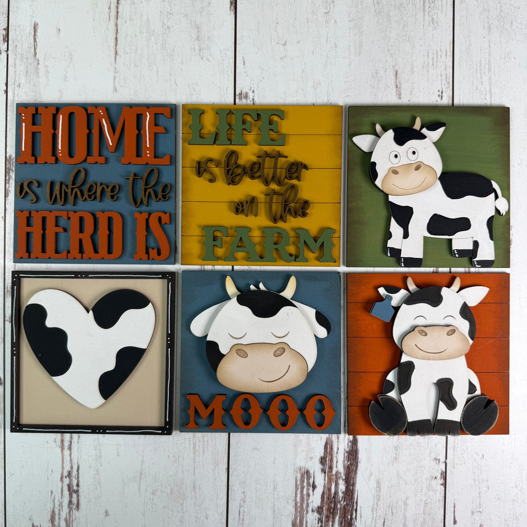 Six rustic decorative wall signs arranged in a 2x3 grid on a whitewashed wood background. The signs feature cow illustrations, farm-themed phrases like 'HOME is where the HERD IS' and 'LIFE is better on the FARM', a heart-shaped cow graphic, and the word 'MOOO'.