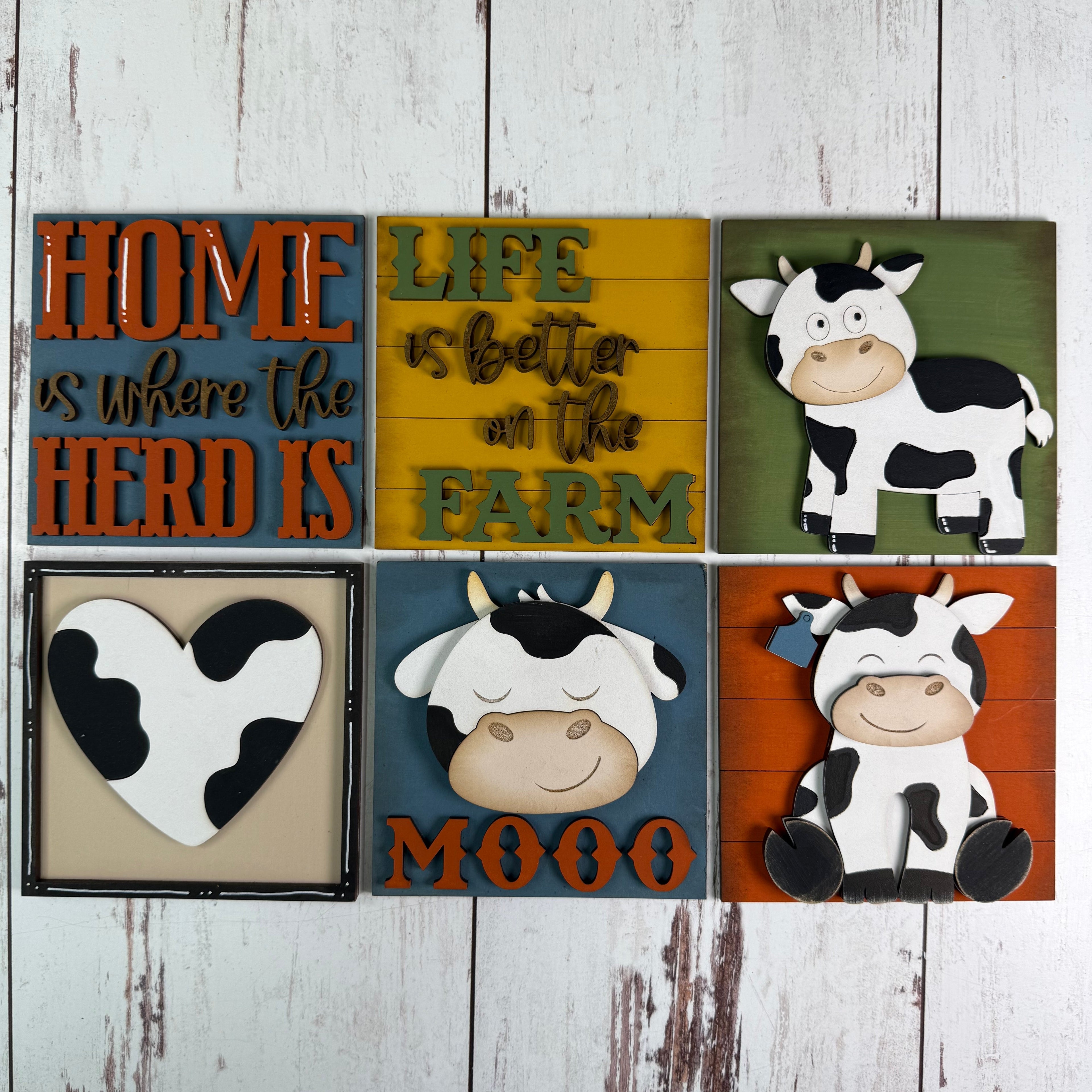 Six rustic decorative wall signs arranged in a 2x3 grid on a whitewashed wood background. The signs feature cow illustrations, farm-themed phrases like 'HOME is where the HERD IS' and 'LIFE is better on the FARM', a heart-shaped cow graphic, and the word 'MOOO'.