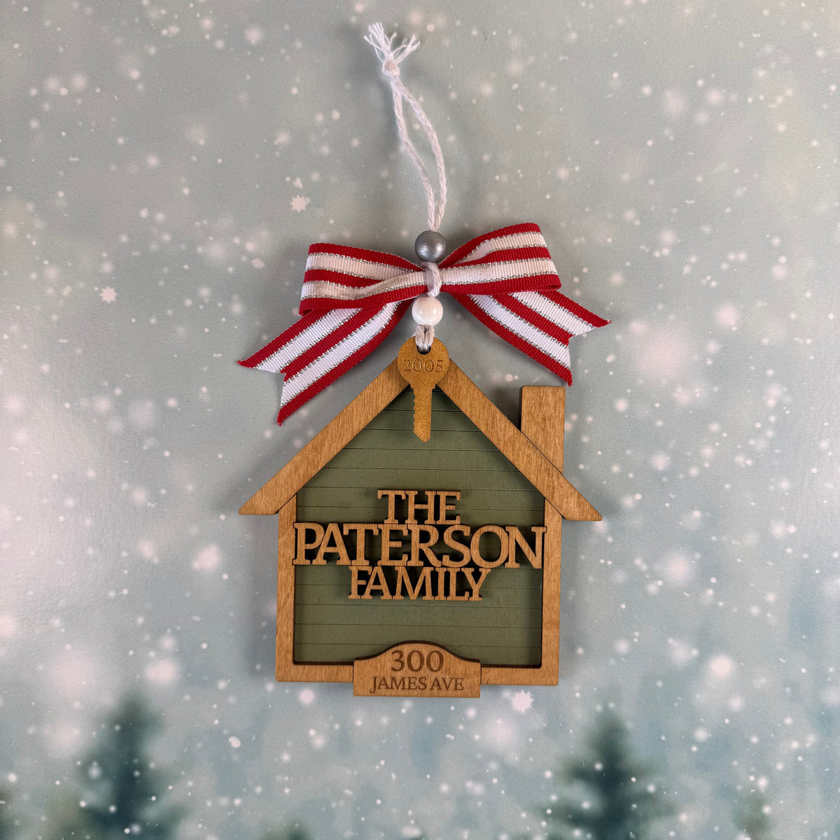 A personalized wooden house-shaped ornament hanging from a red and white striped ribbon. The ornament has a dark green center panel with the text 'THE PATERSON FAMILY' and '300 JAMES AVE' in light wood.