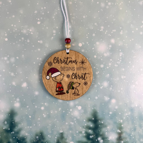 A round wooden Christmas ornament hanging from a white cord with red and gold beads. The ornament features a laser-engraved illustration of a child in a Santa hat and Snoopy with the text 'Christmas BEGINS WITH Christ' against a snowy, blurred winter background.