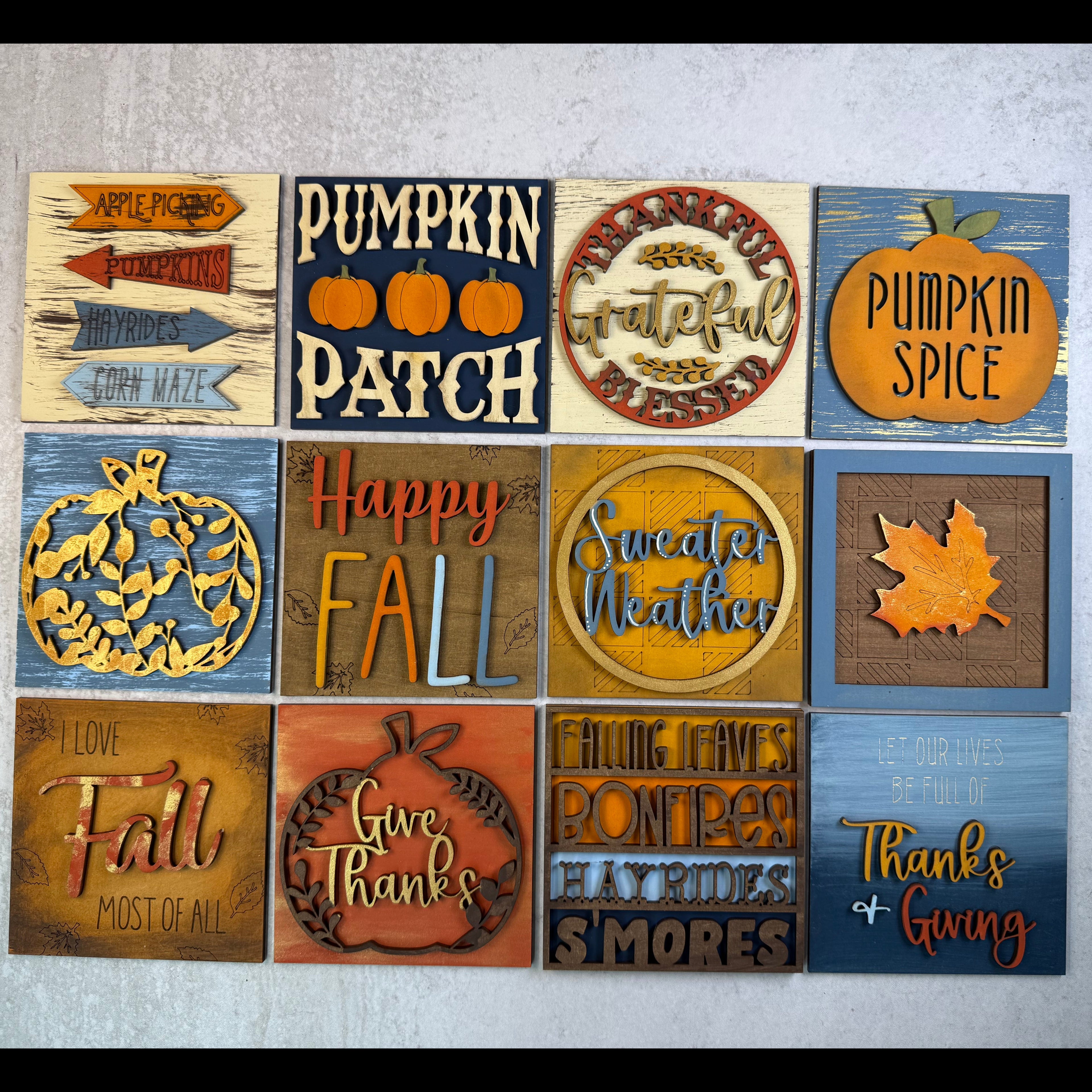A set of 12 square autumn-themed wooden sign plaques arranged in a 3x4 grid on a light gray textured surface. The signs feature various fall designs including pumpkins, leaves, harvest motifs, and seasonal phrases like 'Fall is here', 'Happy Fall', 'Pumpkin Patch', and 'Pumpkin Spice'.