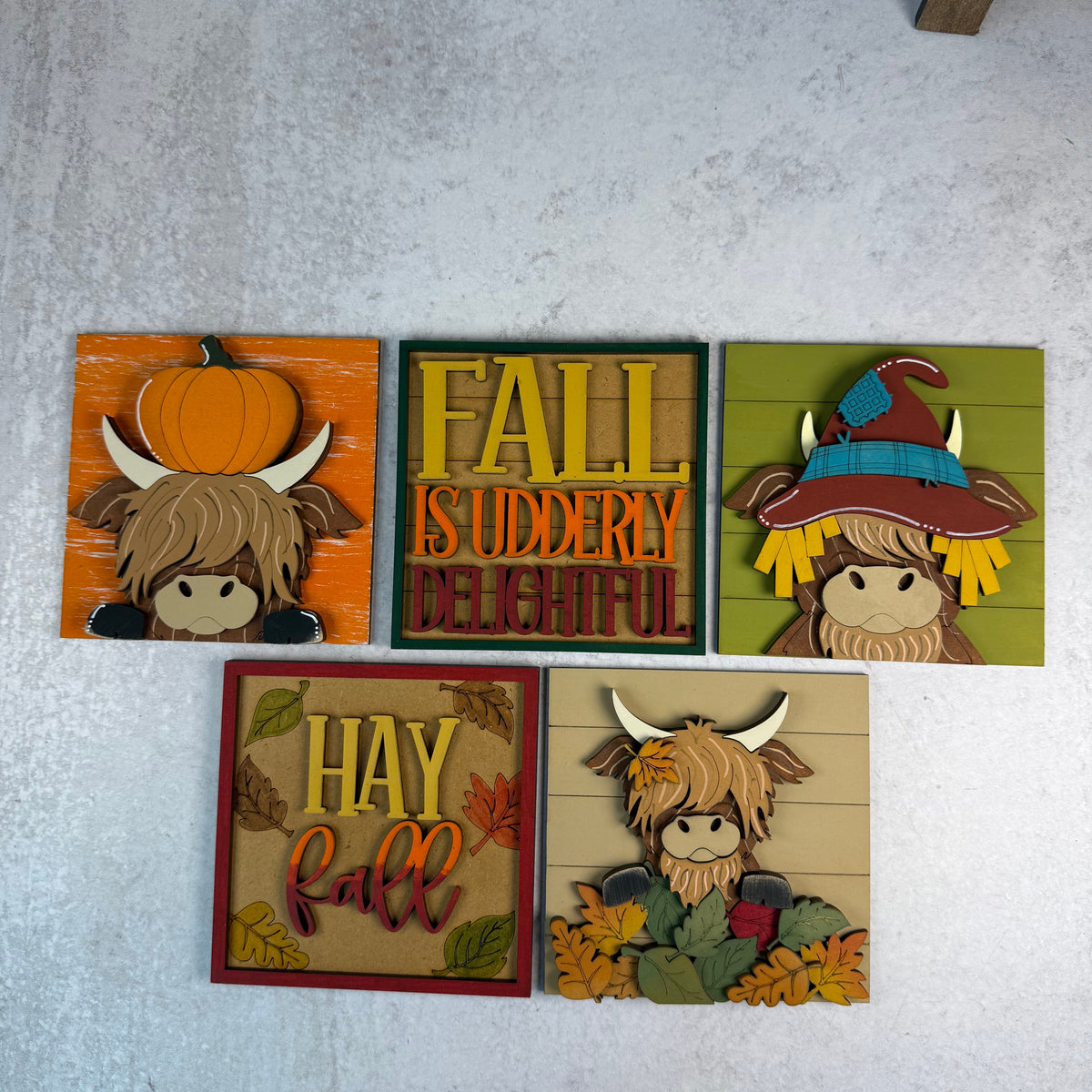 Five autumn-themed wooden plaque signs arranged on a light gray textured surface. The signs feature Highland cows, pumpkins, leaves, and fall quotes including 'FALL IS UDDERLY DELIGHTFUL' and 'HAY FALL'.