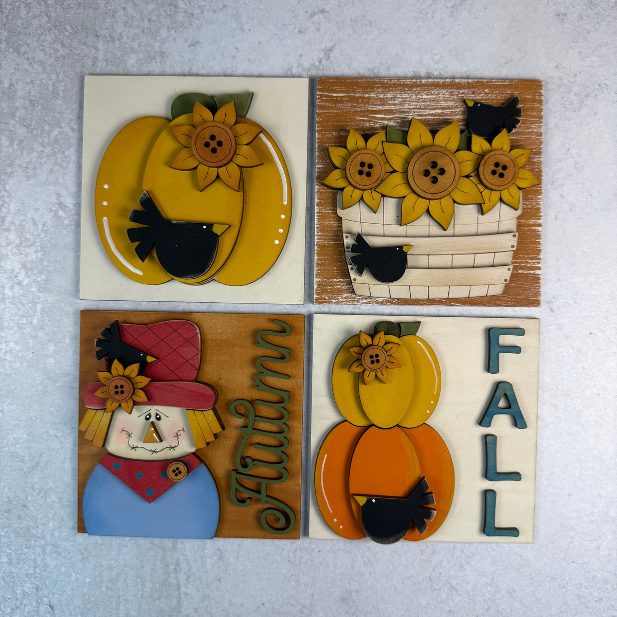 Four square autumn-themed decorative wall plaques arranged in a 2x2 grid on a light gray textured background. The designs include a yellow pumpkin with sunflowers and a black bird, sunflowers in a basket with birds, a scarecrow with the word 'Autumn' and a red hat, and a pumpkin with the word 'FALL' in blue letters.