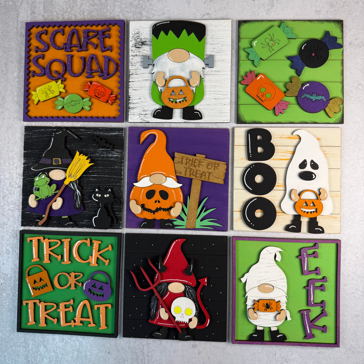 A set of nine square Halloween-themed decorative tiles or signs arranged in a 3x3 grid on a light gray textured background. Each tile features a different spooky design, including pumpkins, ghosts, witches, gnomes, candy, and words like 'Scare Squad,' 'Trick or Treat,' 'Boo,' and 'Eek.