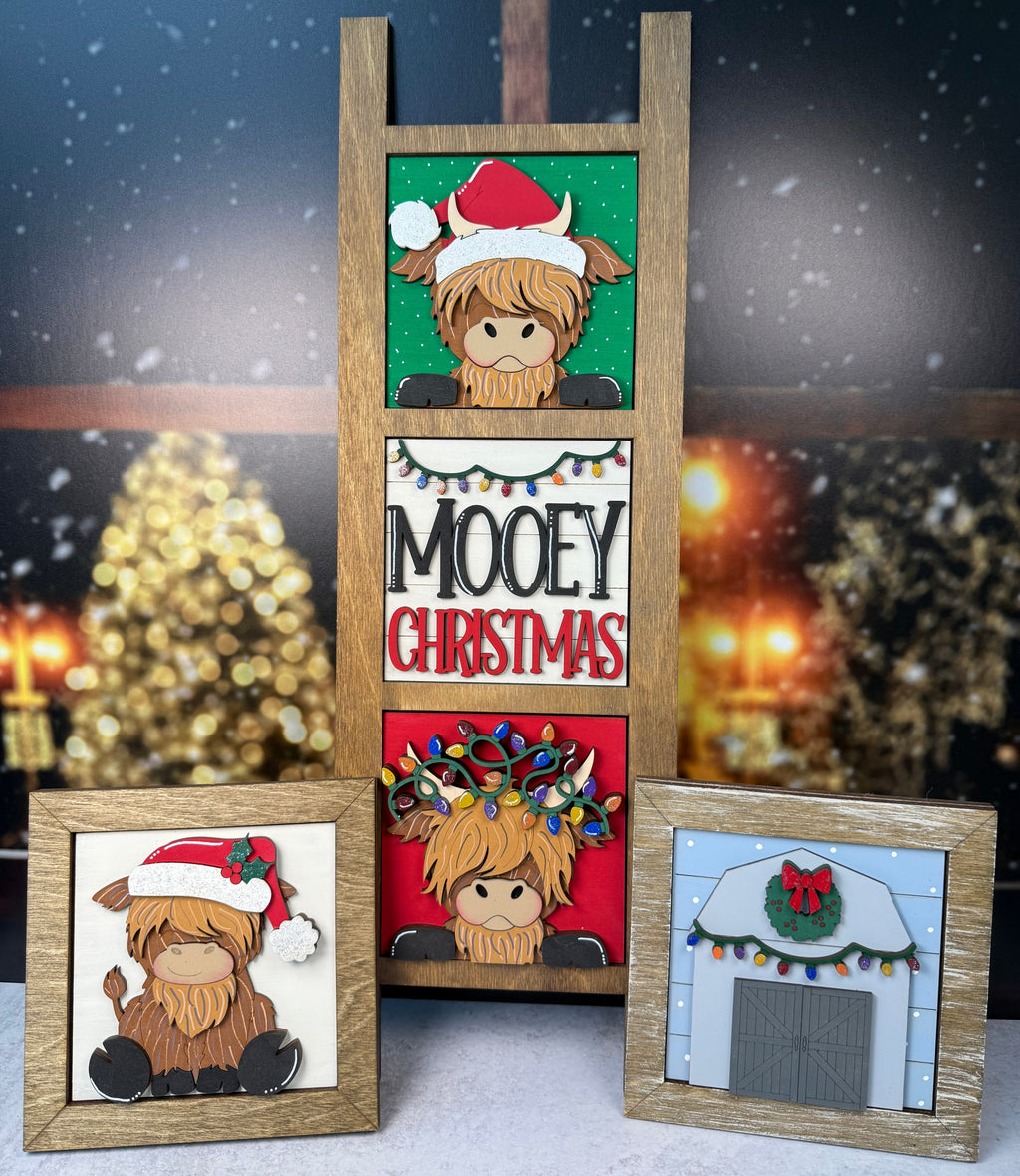 Three handmade wooden Christmas plaque designs displayed together against a festive blurred background of Christmas trees and snow. The center plaque features a wooden ladder frame with three stacked panels showing a Highland cow in a Santa hat on green, white, and red backgrounds with the text 'MOOY CHRISTMAS'.