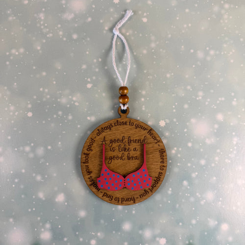 A round wooden hanging ornament with a white cord, featuring an engraved quote and a pink polka-dot bra illustration, shown against a snowy winter background.