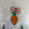 A Christmas ornament shaped like a light bulb, featuring a green top, an orange body with printed holiday poem text, and a red, green, and white striped bow with a white hanging string. The ornament is centered against a soft, blurred winter background with falling snow and dark pine tree silhouettes.