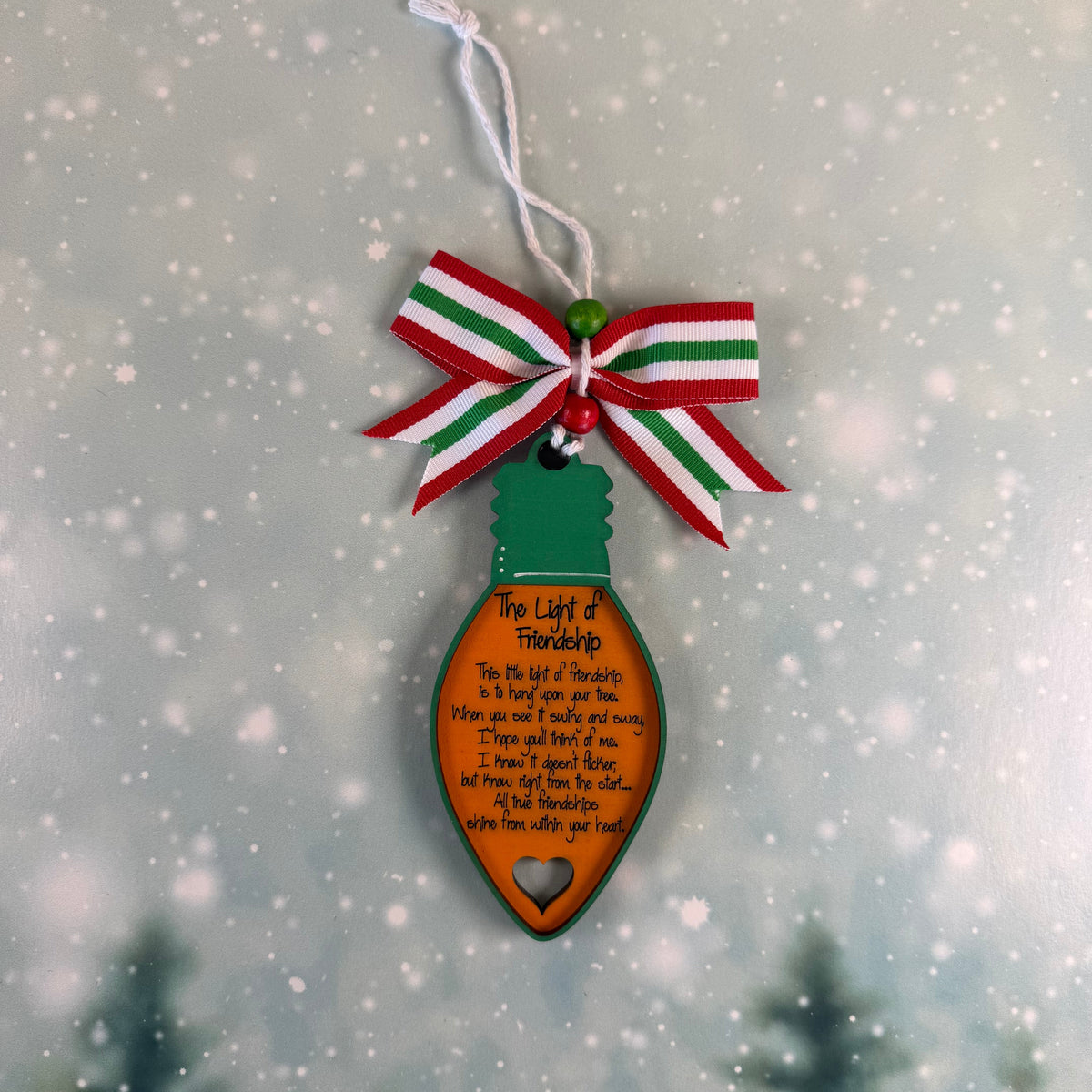 A Christmas ornament shaped like a light bulb, featuring a green top, an orange body with printed holiday poem text, and a red, green, and white striped bow with a white hanging string. The ornament is centered against a soft, blurred winter background with falling snow and dark pine tree silhouettes.