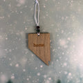 A wooden Nevada-shaped ornament engraved with the word 'home.' hangs from a white cord with dark beads, set against a soft gray background with a snowy bokeh effect.