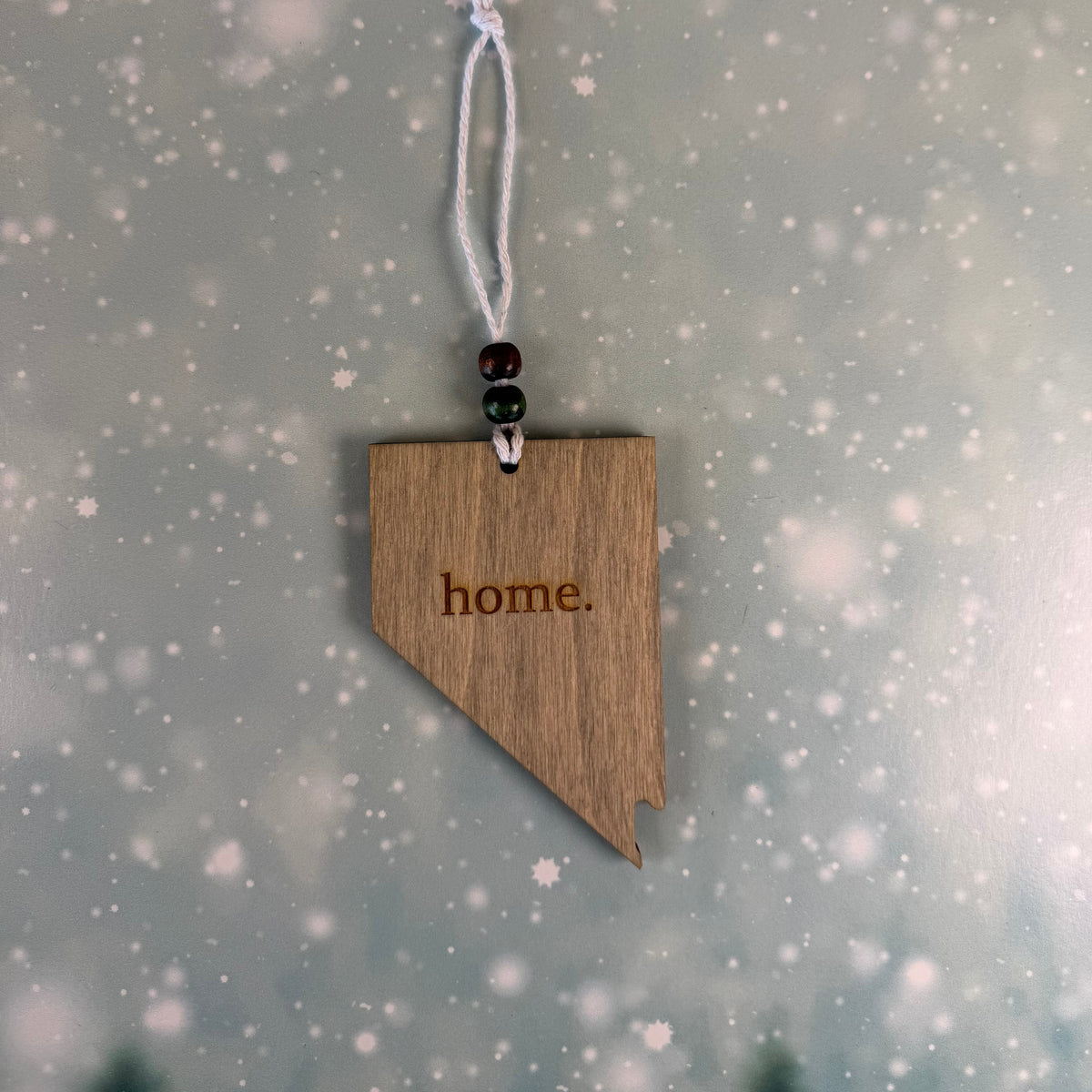 A wooden Nevada-shaped ornament engraved with the word 'home.' hangs from a white cord with dark beads, set against a soft gray background with a snowy bokeh effect.