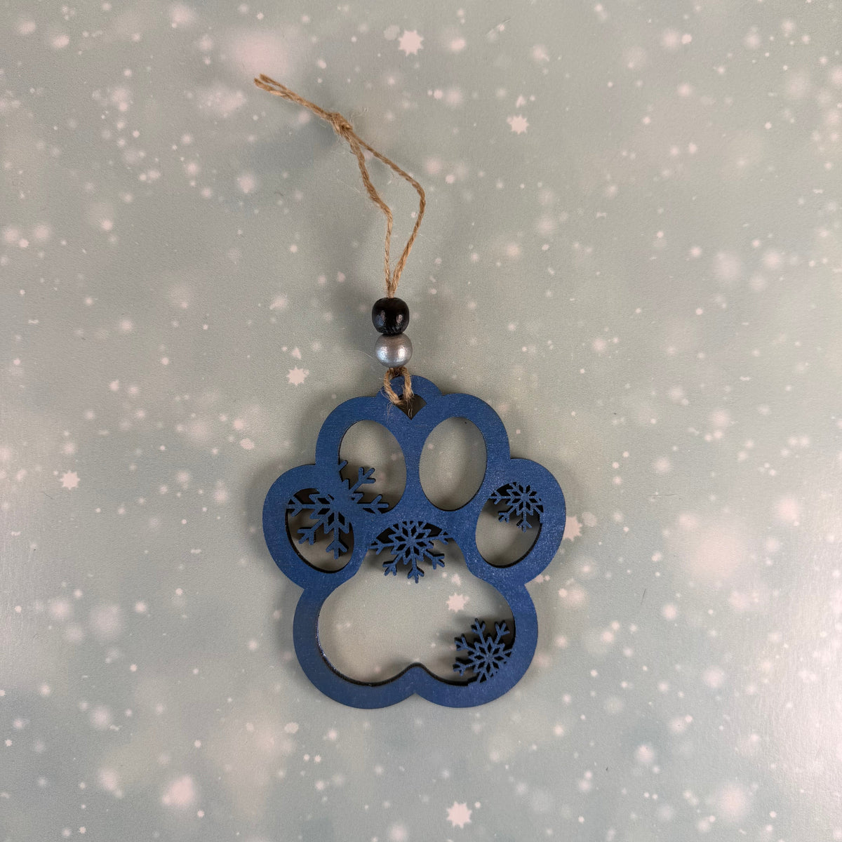 A blue paw print wooden ornament with floral cutout details, hanging from a twine string with a black and white bead, set against a snowy, light blue background.
