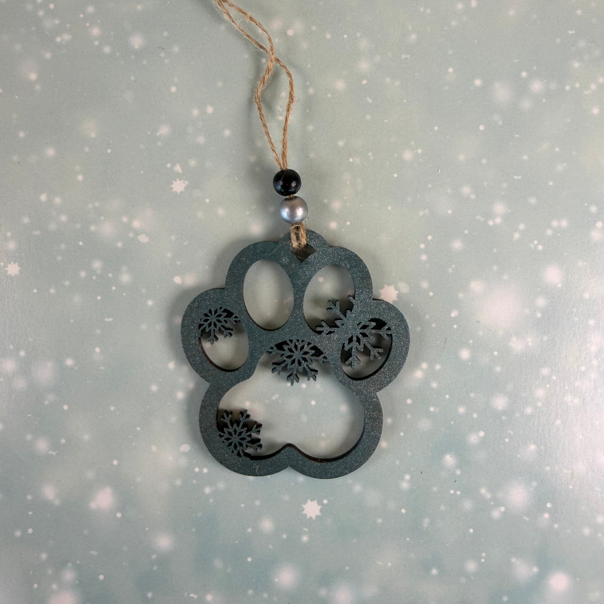 A dark gray wooden paw print ornament with floral cutout details, hanging from a natural jute twine string with black and white beads, set against a soft, snowy blue-green background.